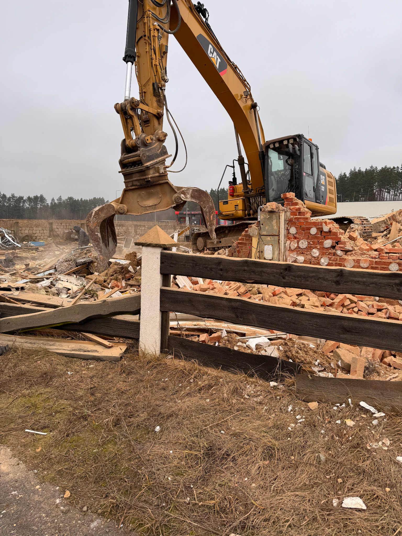 Żółta koparka CAT z chwytakiem rozbiera ceglany budynek. W tle widać gruz i fragmenty konstrukcji. Na pierwszym planie drewniane elementy ogrodzenia.