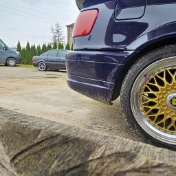 Mobilna wulkanizacja płock 24h7 Paradowski Marcin - Fragment tylnej części granatowego samochodu z widocznym spojlerem i złotą felgą, ujęcie zza drewnianej konstrukcji w pochmurny dzień.