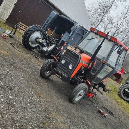 Mobilna wulkanizacja płock 24h7 Paradowski Marcin - Czerwony traktor Ursus 3512 w trakcie naprawy, z częściowo zdemontowanym przednim kołem, stoi na żwirowym podłożu przed budynkiem gospodarczym, obok zaparkowany bus z otwartymi drzwiami, w którym...