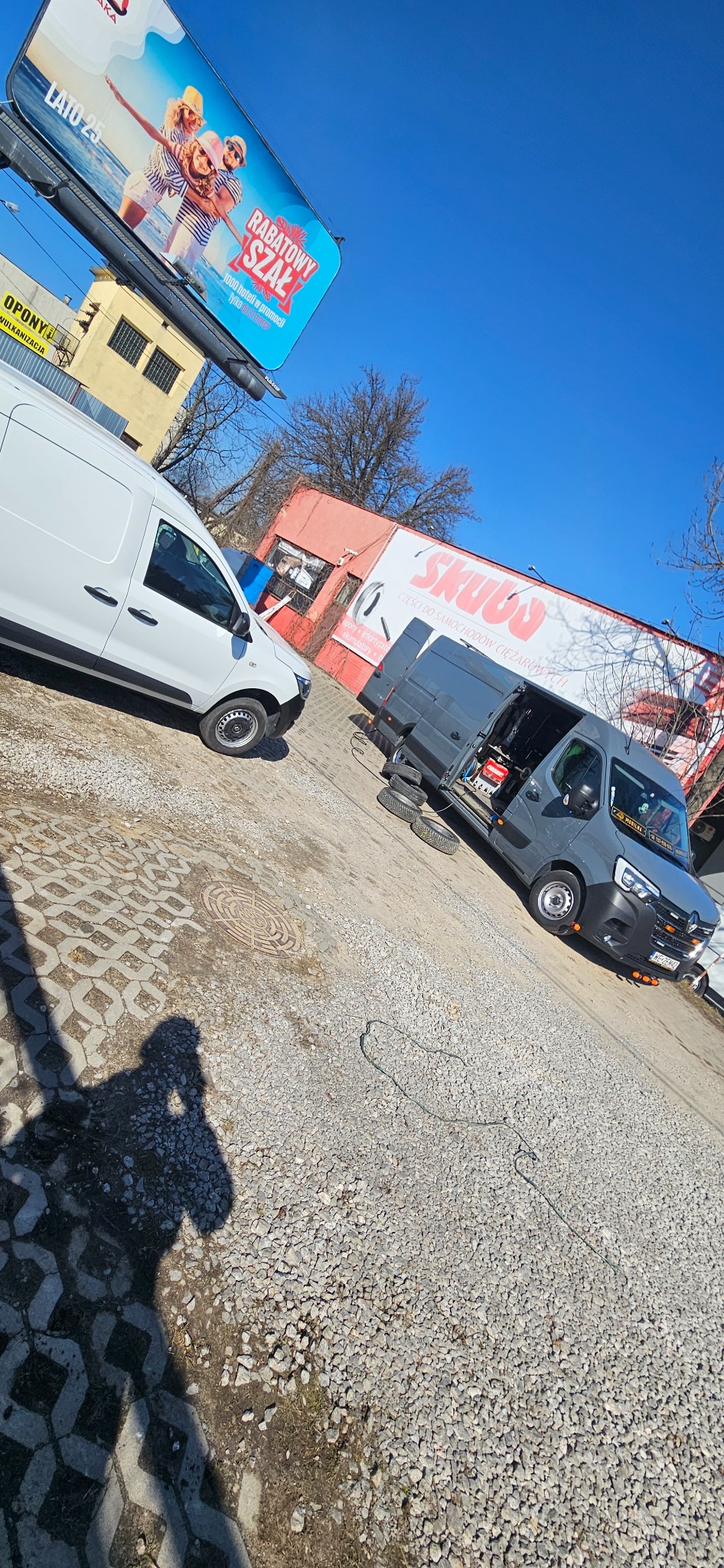 Dwa busy zaparkowane na żwirowym placu przed budynkiem z szyldem 'Skuba części do samochodów ciężarowych', w tle billboard reklamowy.