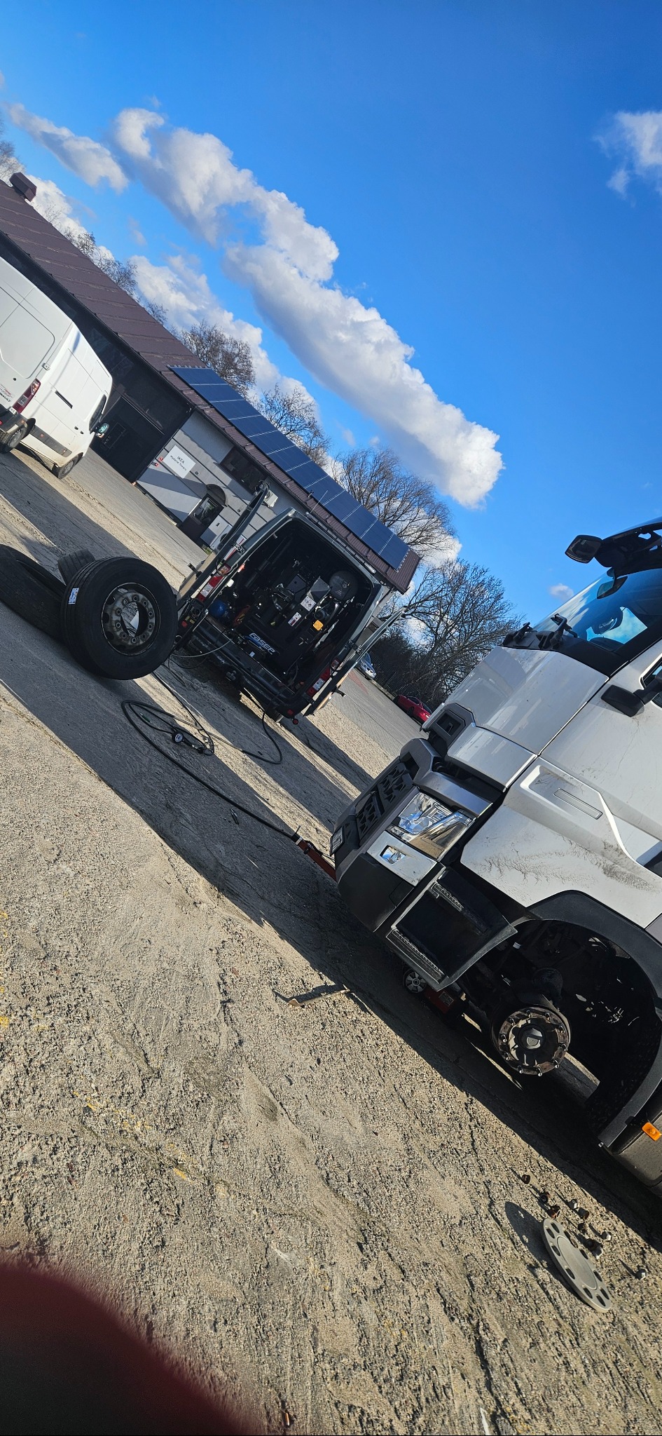 Mobilny serwis ciężarowy na placu: biały ciągnik siodłowy z odkręconym kołem, obok zaparkowany bus serwisowy z otwartymi drzwiami, w tle budynek z panelami słonecznymi.