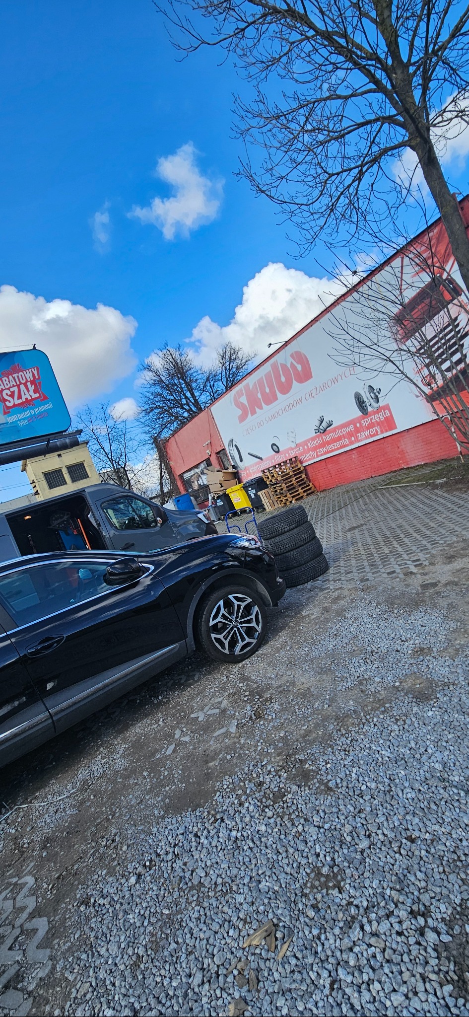 Czarny samochód osobowy i szary van zaparkowane na żwirowym placu przed budynkiem z reklamą części samochodowych, obok stos opon.