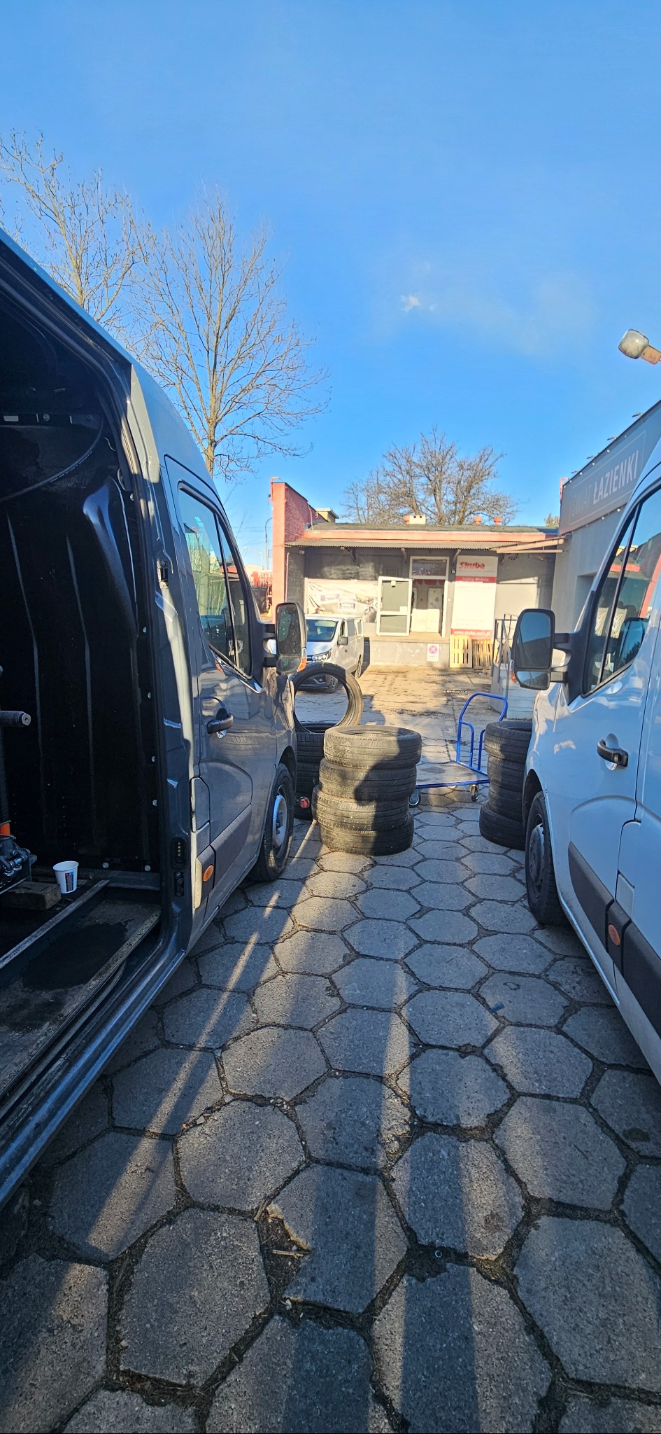 Dwa busy zaparkowane naprzeciwko siebie na brukowanym placu, pomiędzy nimi ułożone w stosy opony, w tle budynek warsztatowy z szyldem.