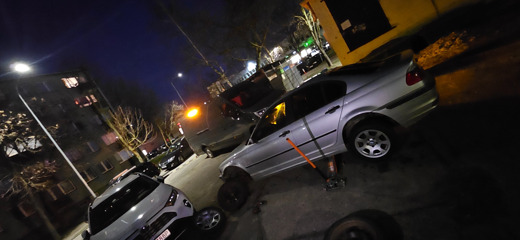 Srebrny samochód osobowy podniesiony na podnośniku hydraulicznym na tle nocnego miejskiego krajobrazu, z widocznymi zdjętymi kołami i pomarańczowym kluczem do kół