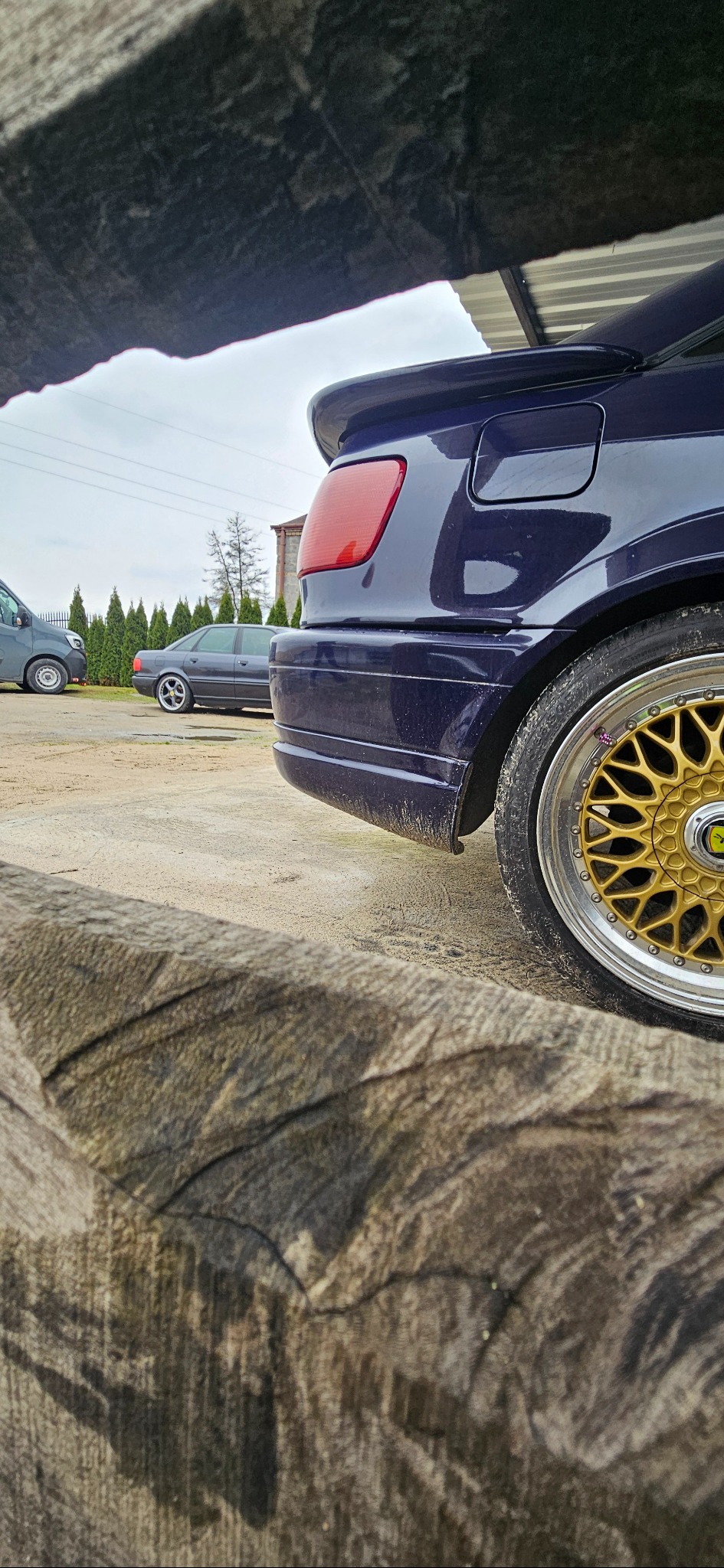 Fragment tylnej części granatowego samochodu z widocznym spojlerem i złotą felgą, ujęcie zza drewnianej konstrukcji w pochmurny dzień.