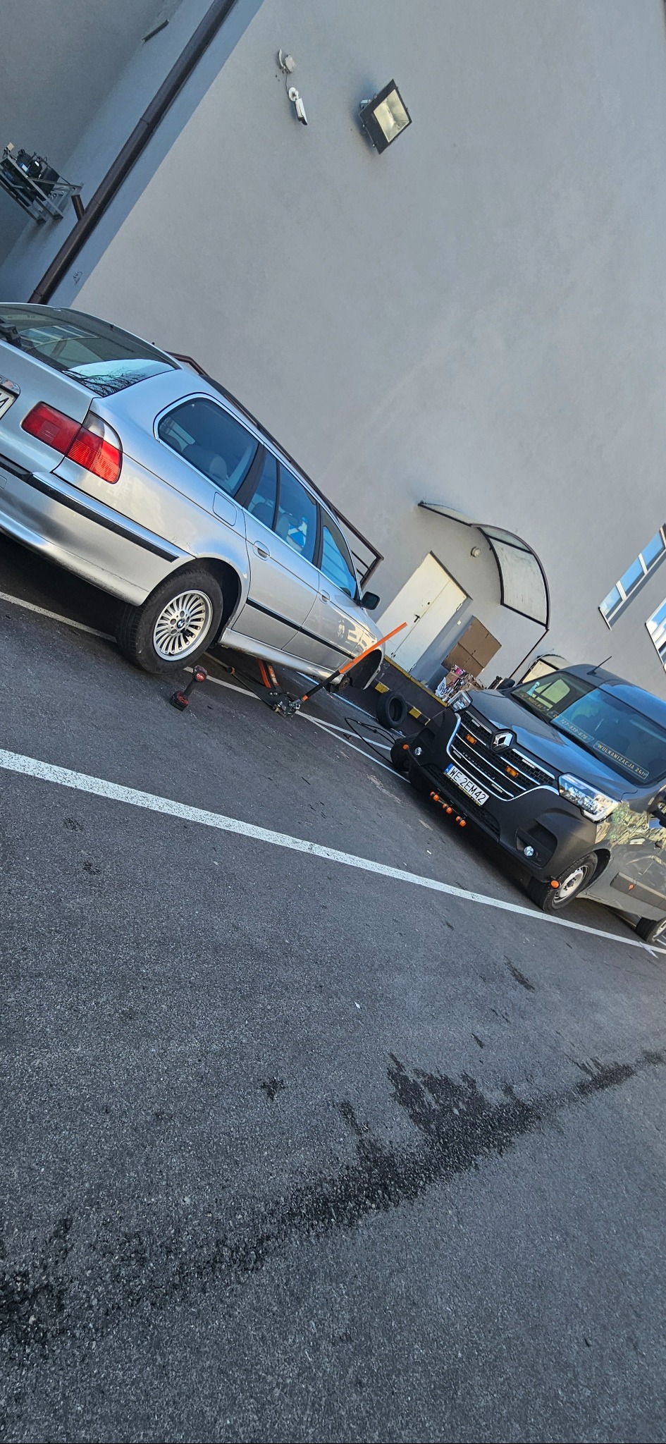 Srebrne kombi BMW na podnośniku hydraulicznym, bez koła, obok czarna laweta Renault z włączonymi pomarańczowymi światłami ostrzegawczymi, na parkingu z widocznymi śladami opon.