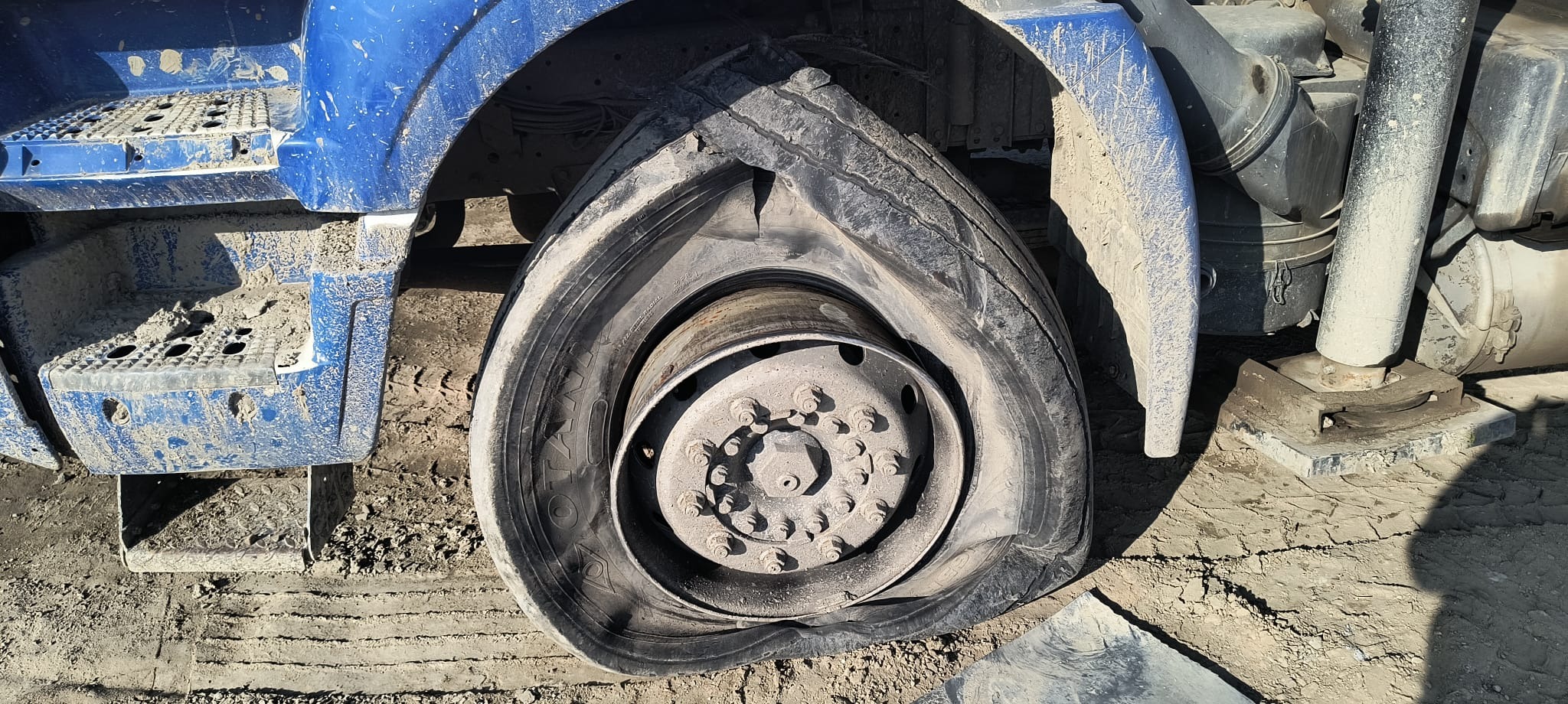 Przebita opona w niebieskiej ciężarówce, widoczne ślady błota na oponie i karoserii, felga z wieloma śrubami