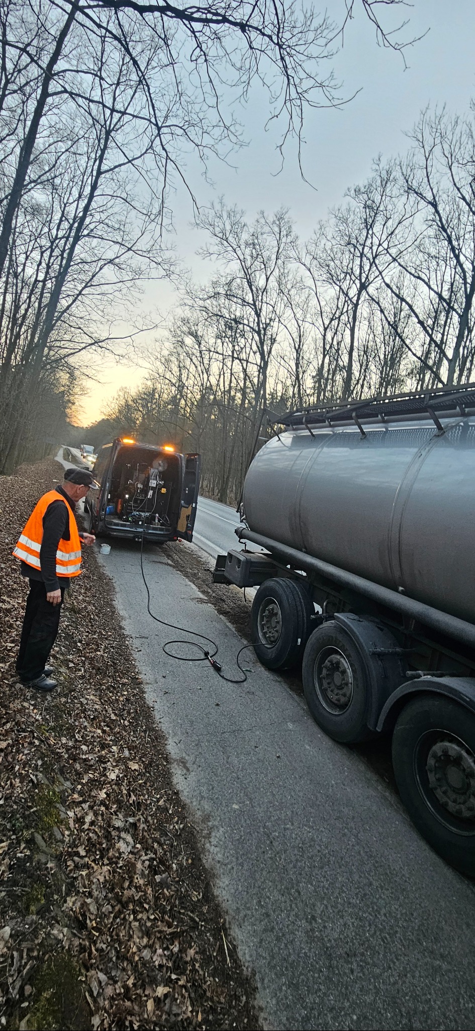 Serwis mobilny przy cysternie: mężczyzna w kamizelce odblaskowej podłącza przewód z agregatu prądotwórczego w busie do instalacji cysterny na poboczu drogi w lesie.