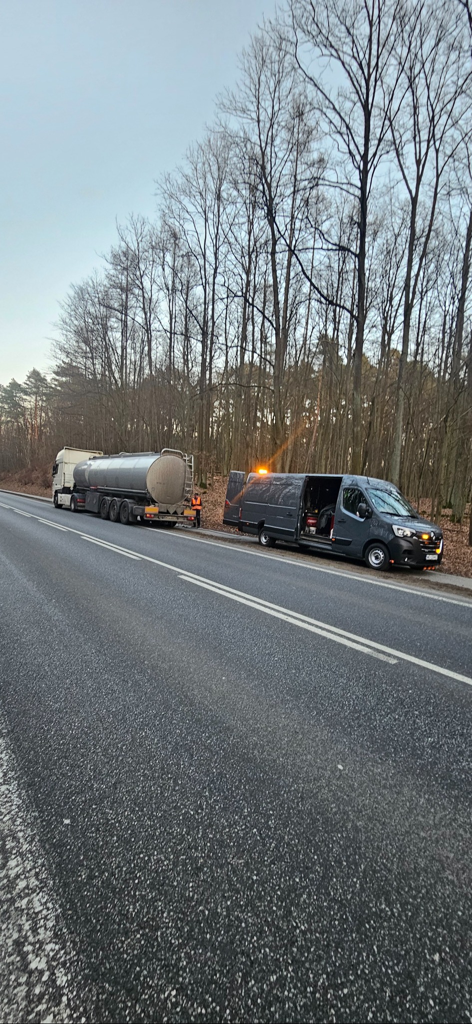 Awaryjne holowanie cysterny na poboczu drogi, z otwartymi drzwiami busa serwisowego i włączonymi światłami ostrzegawczymi, w tle las bez liści.