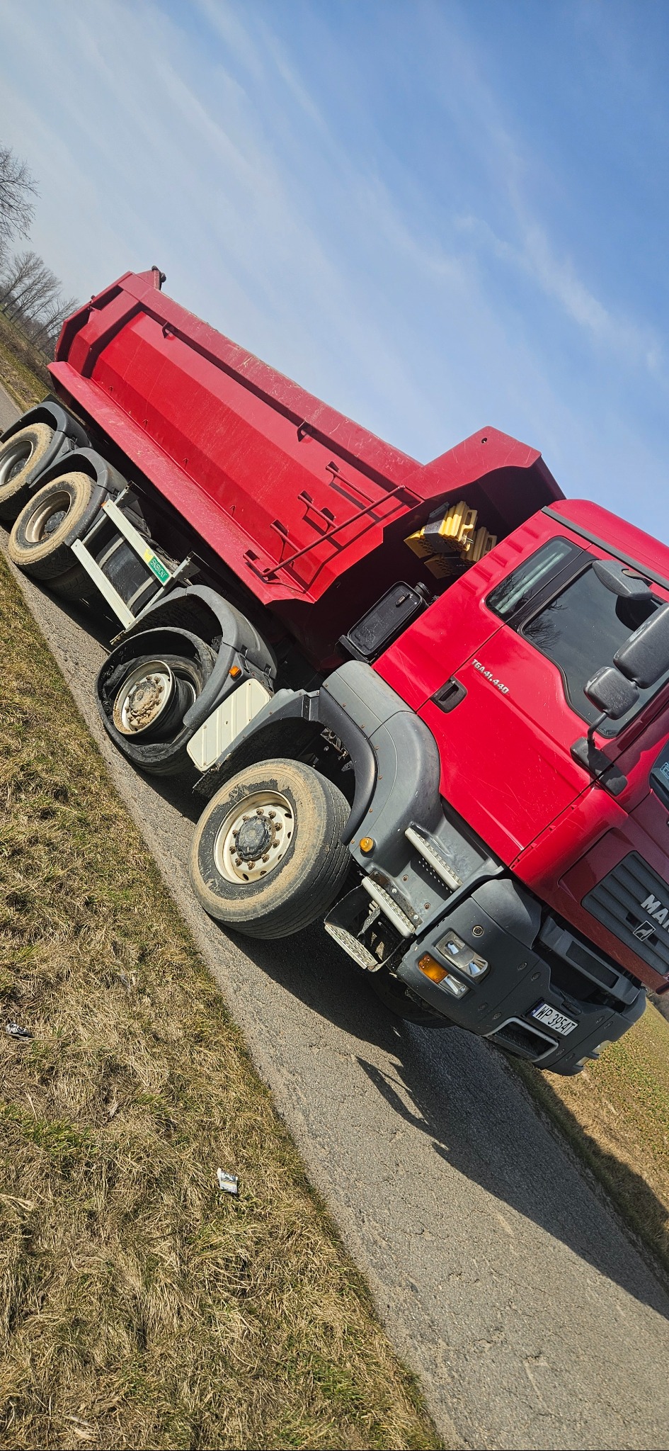 Czerwona wywrotka MAN TGA 41.440 na poboczu drogi, z podniesioną skrzynią i widocznymi tablicami rejestracyjnymi.