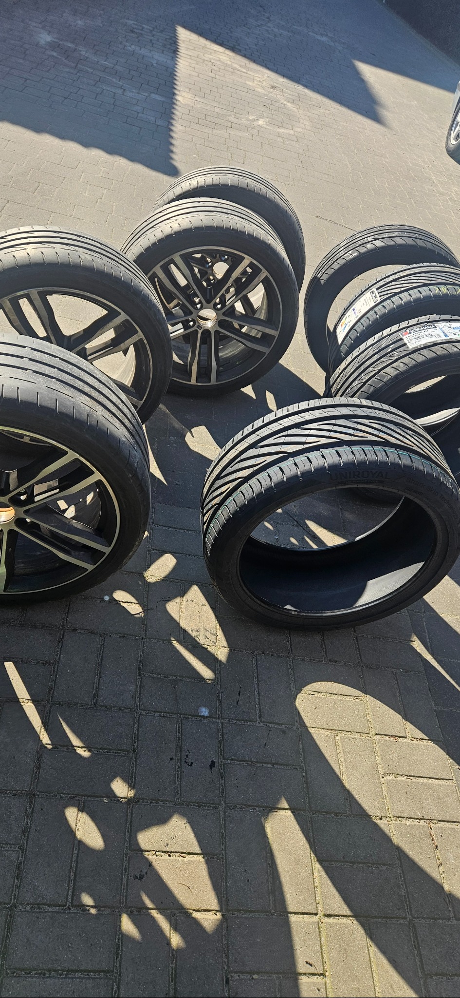 Zestaw nowych opon letnich z felgami aluminiowymi, ułożonych na brukowanej powierzchni w ostrym słońcu, widoczne etykiety producenta na oponach.