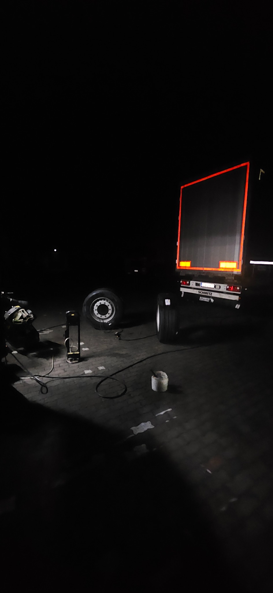 Wymiana opony w nocy przy naczepie ciężarówki, widoczne narzędzia i opony.