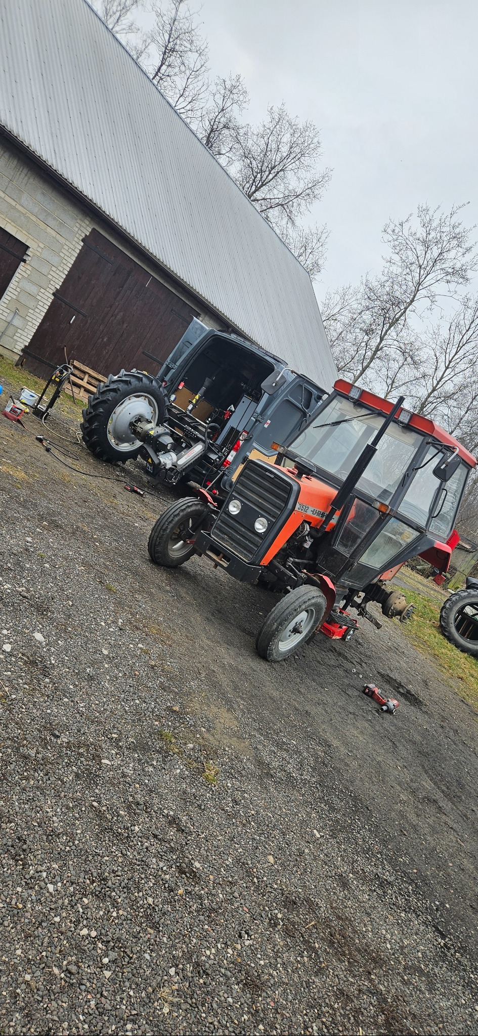 Czerwony traktor Ursus 3512 w trakcie naprawy, z częściowo zdemontowanym przednim kołem, stoi na żwirowym podłożu przed budynkiem gospodarczym, obok zaparkowany bus z otwartymi drzwiami, w którym...