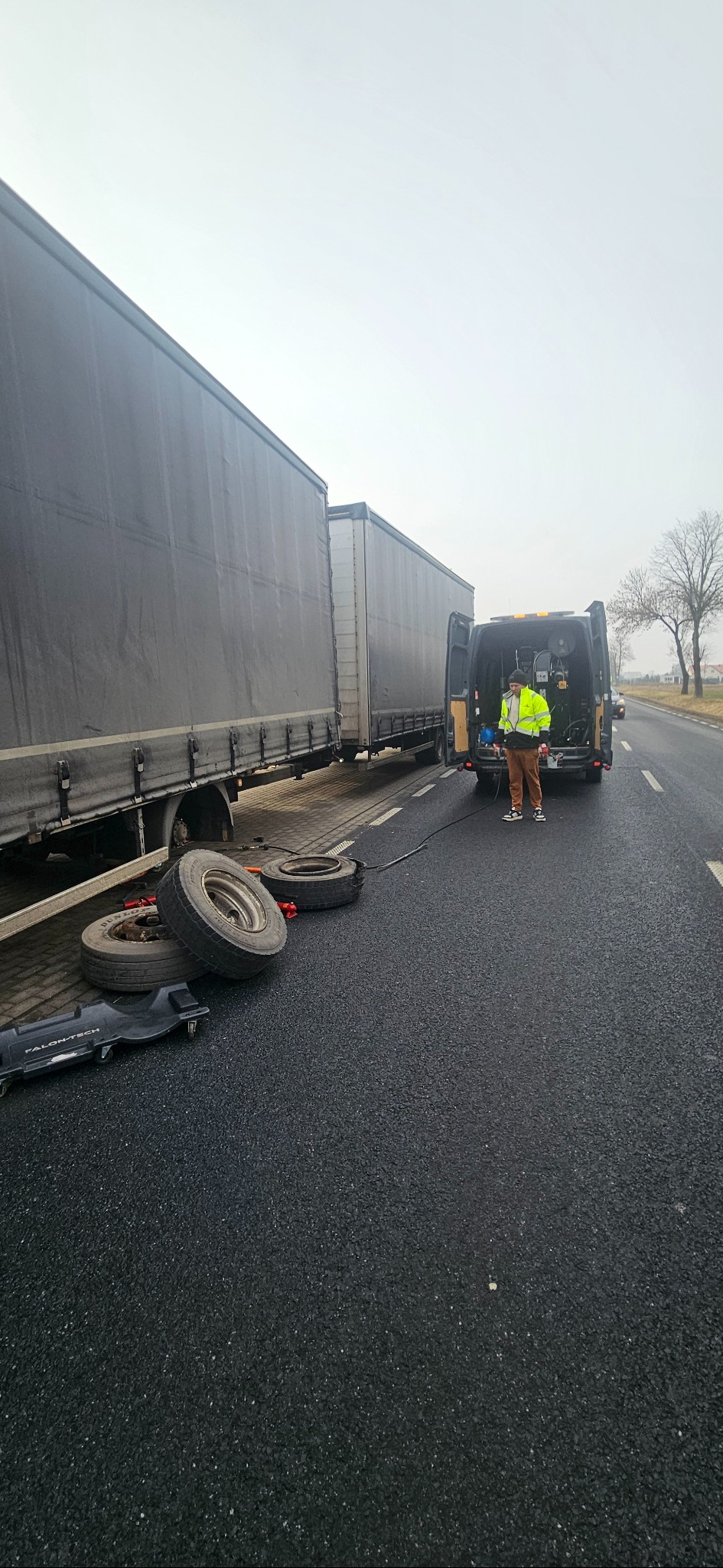 Awaryjna wymiana opon w naczepie ciężarowej na poboczu drogi, z użyciem mobilnego serwisu z samochodu dostawczego; widoczne opony i narzędzia na asfalcie.