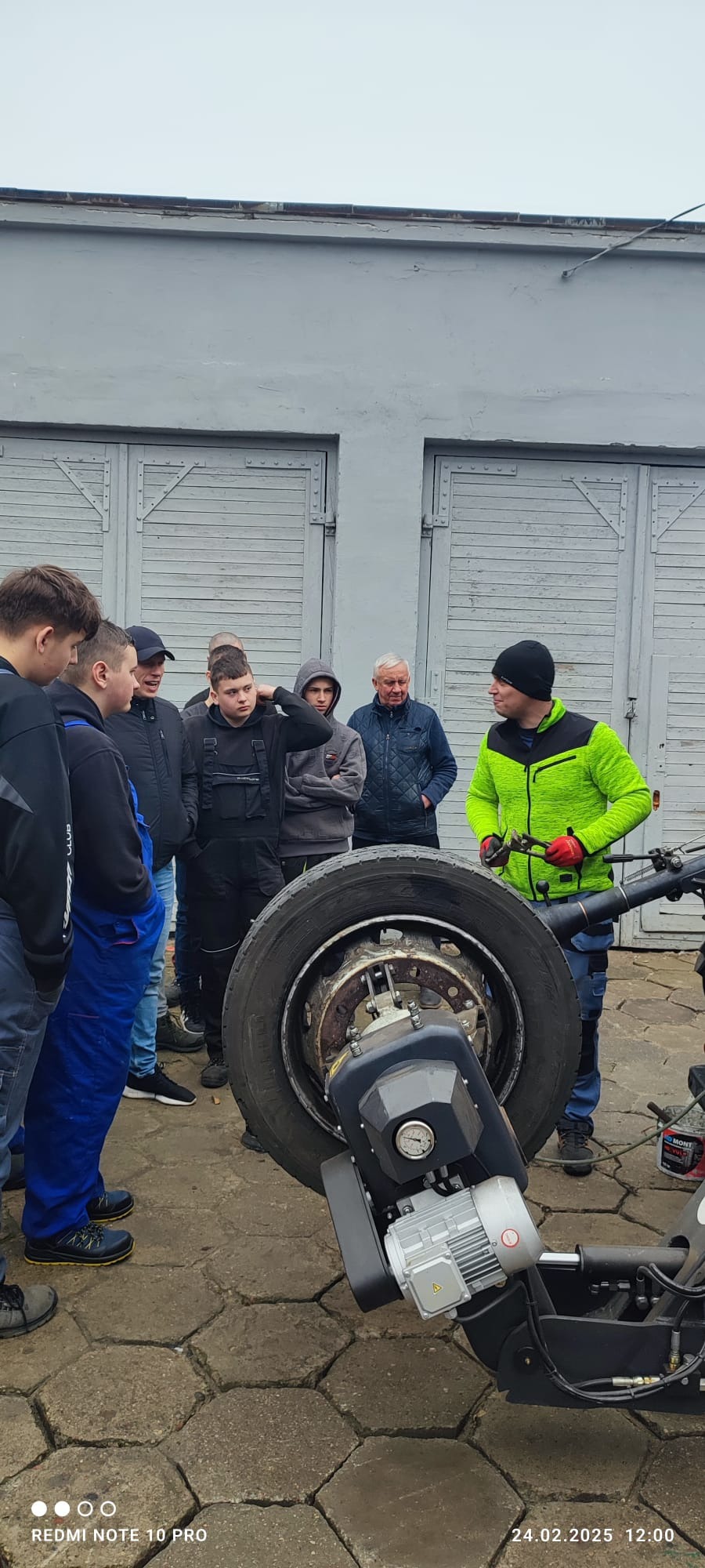 Grupa osób obserwuje demontaż opony ciężarowej przy użyciu specjalistycznego urządzenia w warsztacie, widoczne narzędzia i elementy pojazdu.