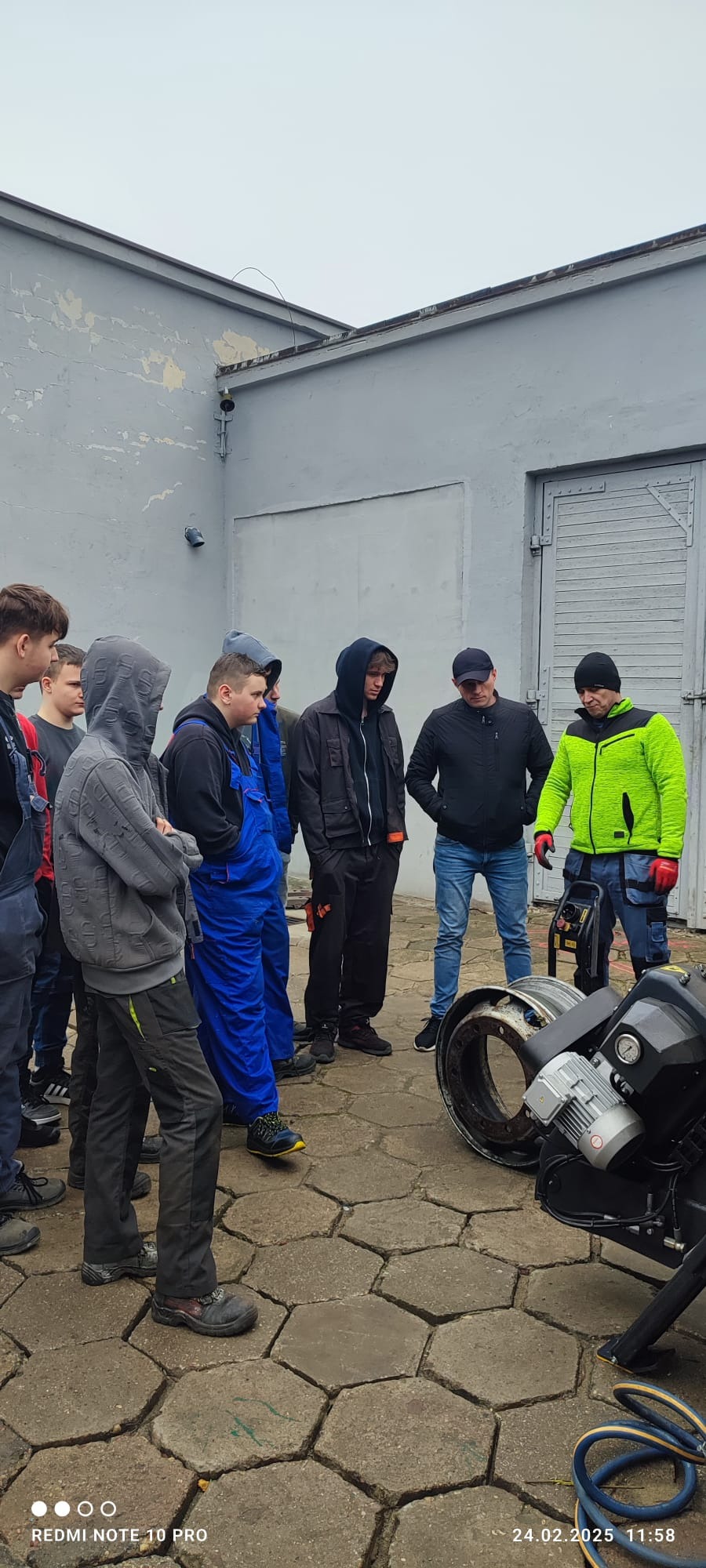 Grupa osób, w tym jeden w niebieskim kombinezonie roboczym i jeden w jaskrawozielonej kurtce, zgromadzona wokół urządzenia do demontażu opon ciężarowych na brukowanym placu przed szarym budynkiem.