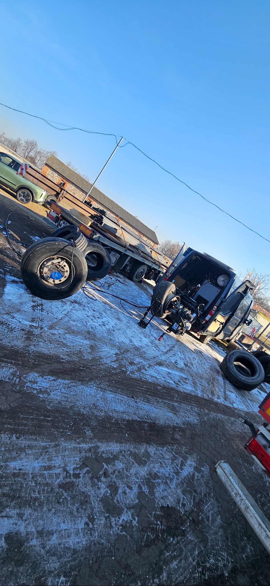 Częściowo rozebrana ciężarówka na placu z oponami obok, widoczne elementy podwozia i ramy, w tle budynek i zaparkowany samochód osobowy.