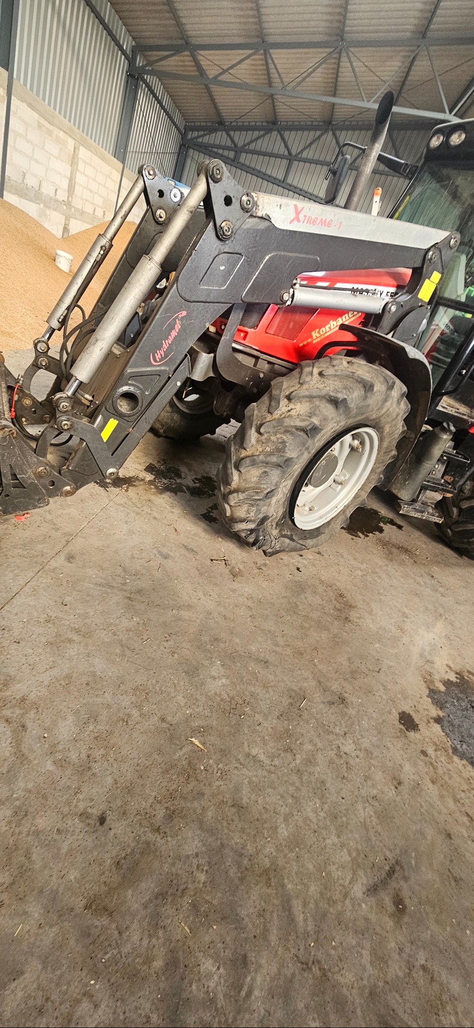 Czerwony ciągnik rolniczy z szarym ładowaczem czołowym Xtreme-1 wewnątrz magazynu z widoczną kupą ziarna w tle.