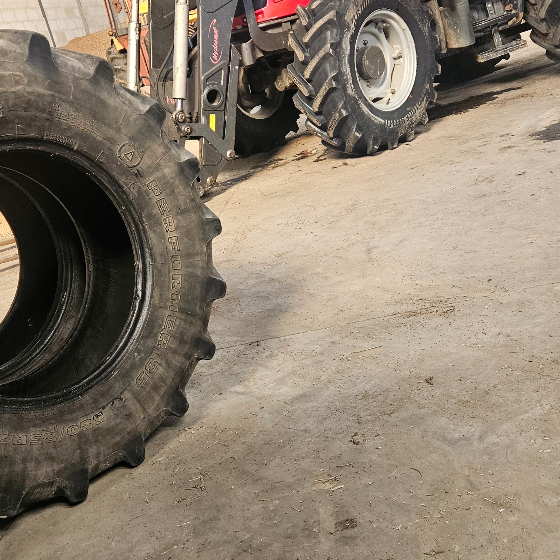 Dwie opony do ciągnika rolniczego, jedna leży na posadzce, druga zamontowana na ciągniku z podnośnikiem hydraulicznym, widoczne oznaczenia 380/85R28 Performer 85.