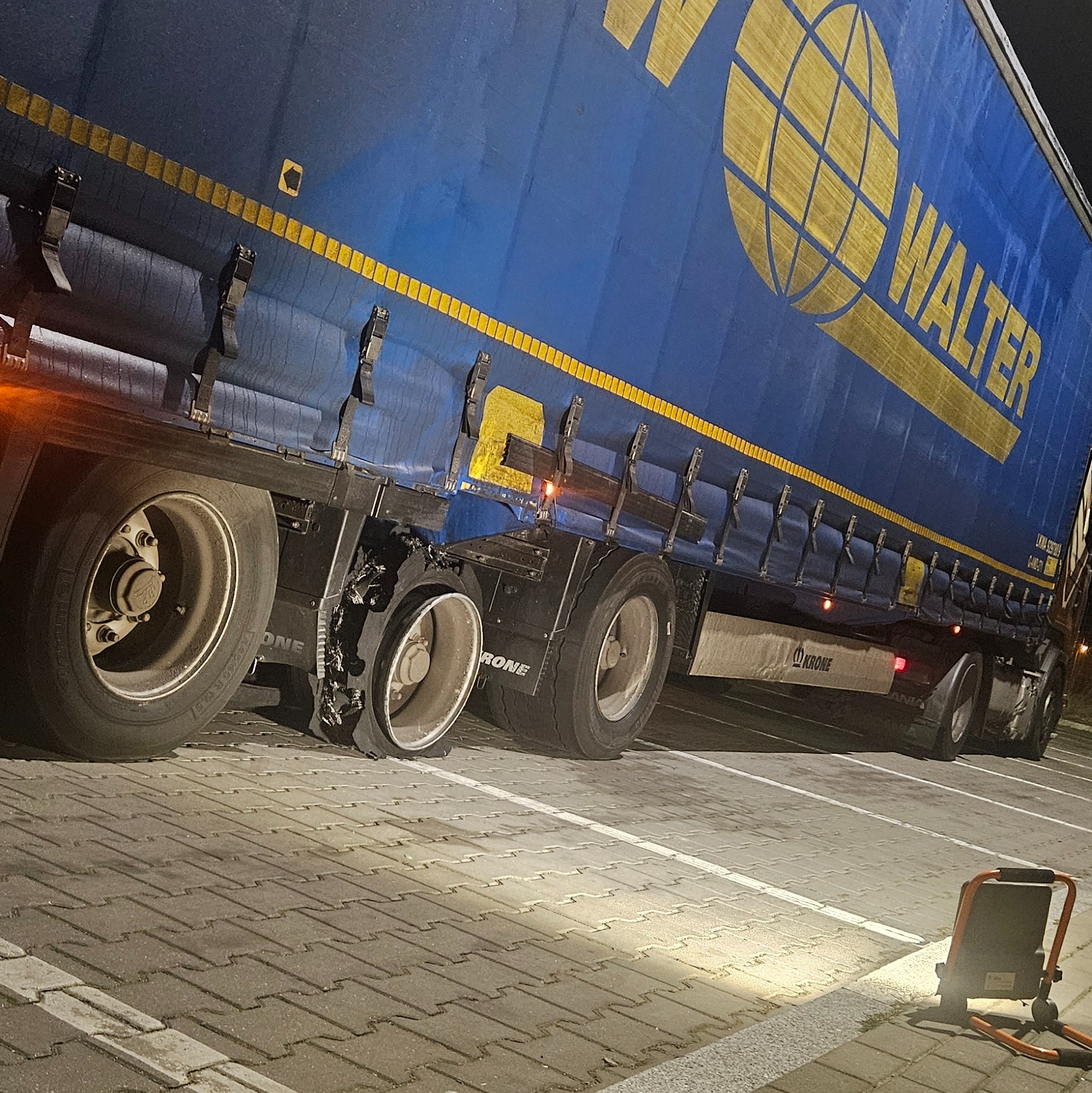 Niebieska naczepa ciężarówki z logo WALTER i uszkodzoną oponą, stojąca na parkingu oświetlonym lampą roboczą.