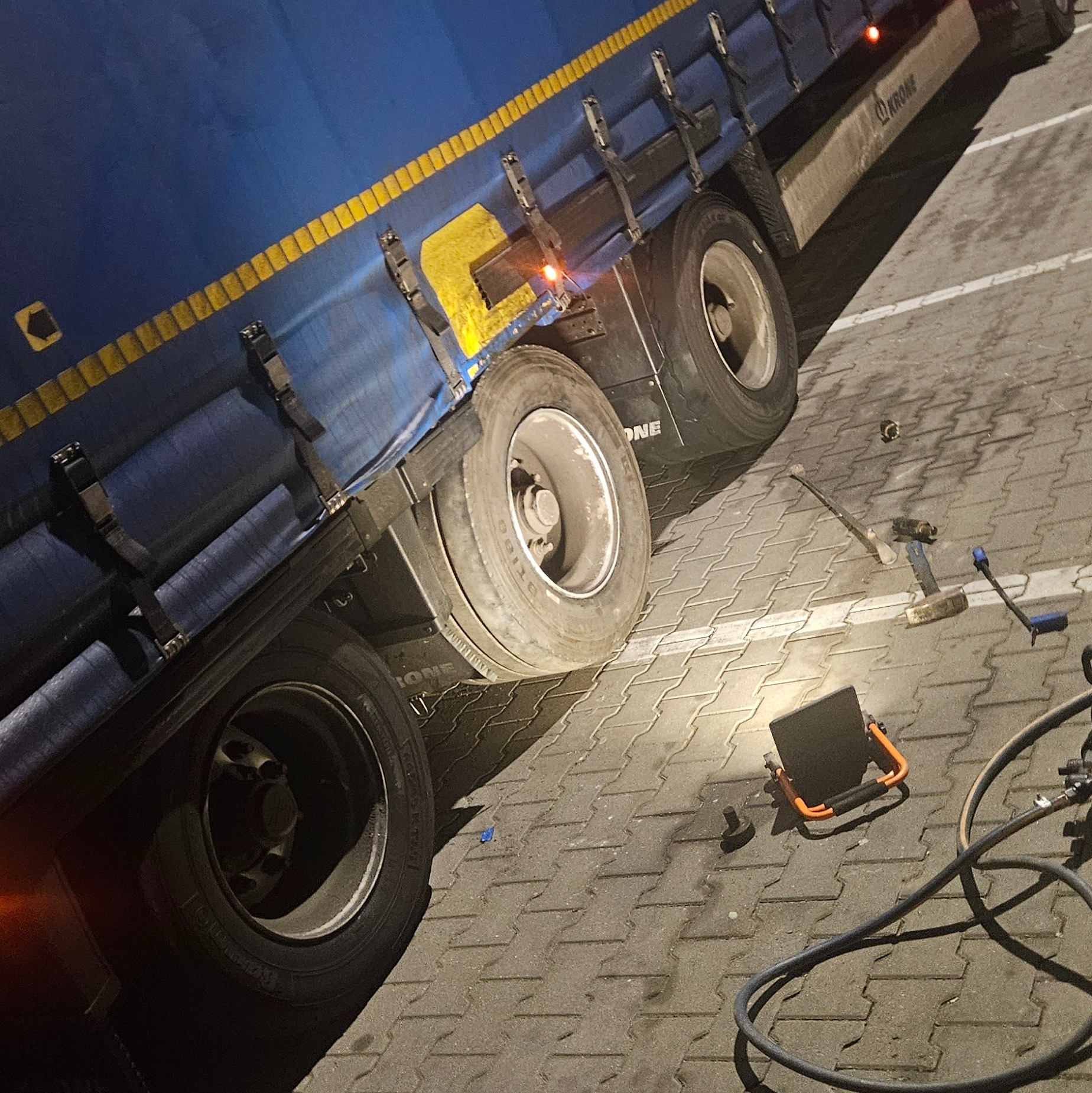 Ciężarówka z niebieską plandeką i żółtymi pasami, widoczne koła, narzędzia leżące na ziemi, lampa robocza oświetla obszar naprawy.