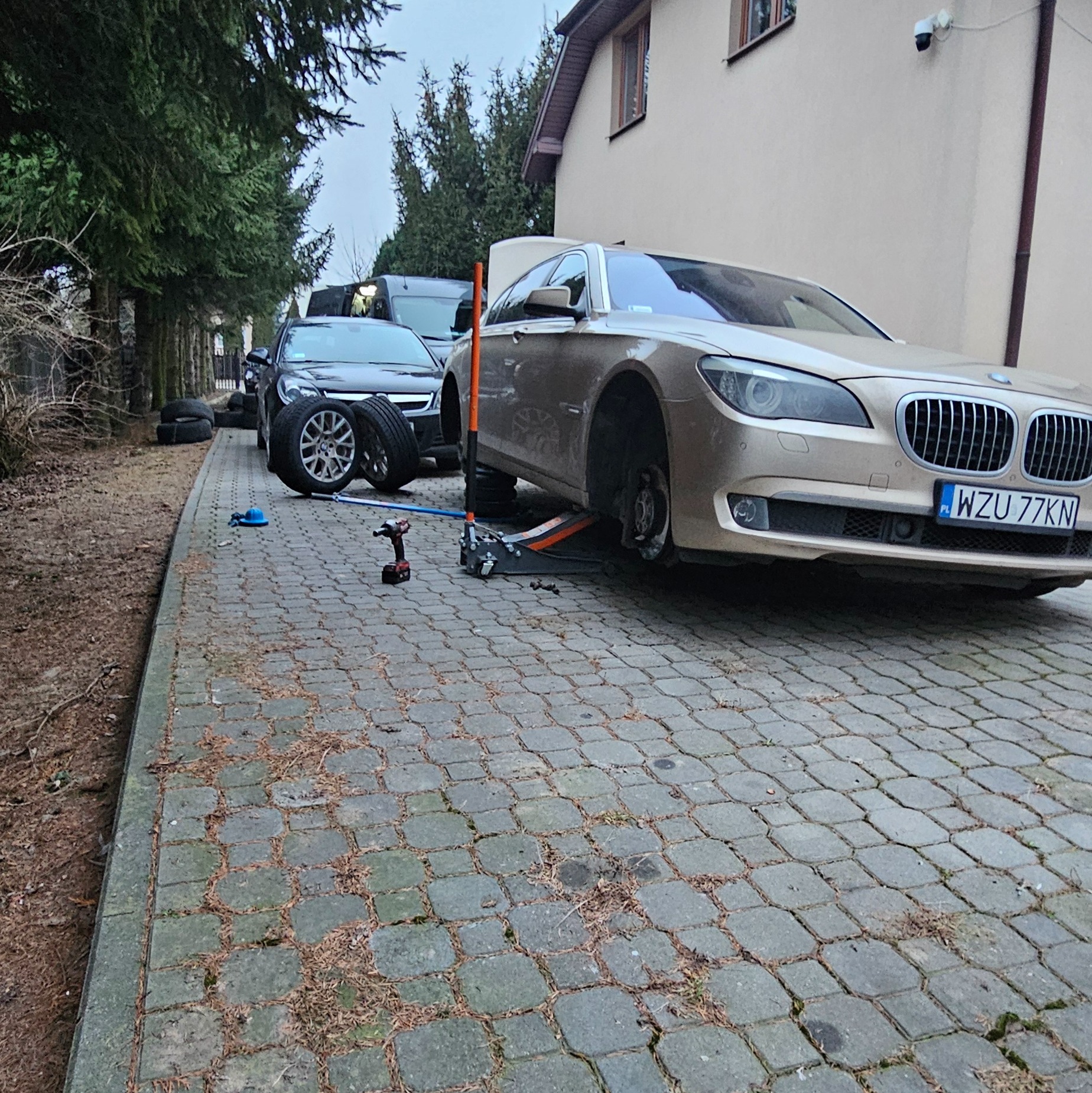 Złoty samochód BMW serii 7 na podnośniku hydraulicznym z odkręconym kołem na brukowanym podjeździe, w tle zaparkowane inne pojazdy i opony, widoczny klucz pneumatyczny i narzędzia.