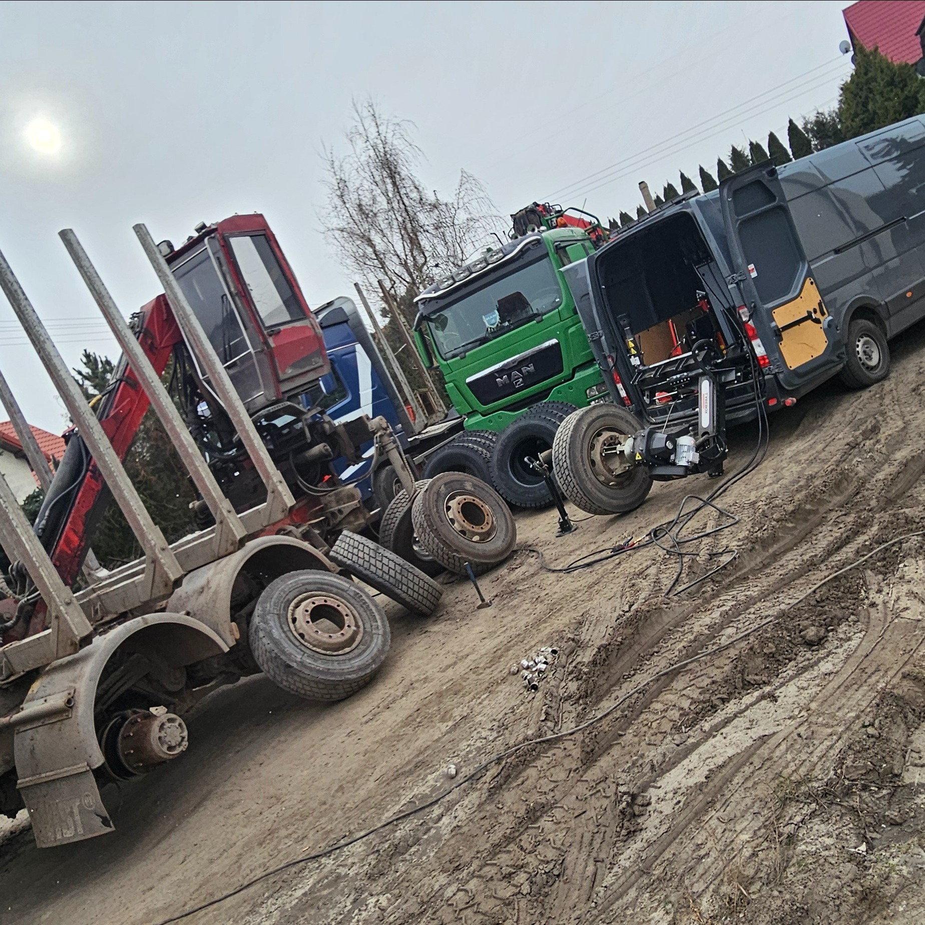 Ciężarówki, w tym MAN, oraz przyczepa do transportu drewna na placu, zdemontowane koła i serwisowy bus z urządzeniem do wymiany opon.