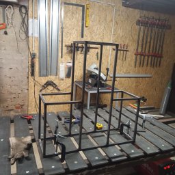 Steel and wood - Konstrukcja metalowa w trakcie spawania na stole warsztatowym, z narzędziami i spawarką w tle.