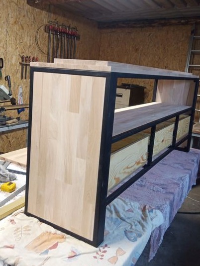 Szafka RTV w trakcie montażu: surowe drewno i czarna metalowa rama, widoczne niedociągnięcia wykończeniowe, w tle warsztat stolarski z klamrami i narzędziami.