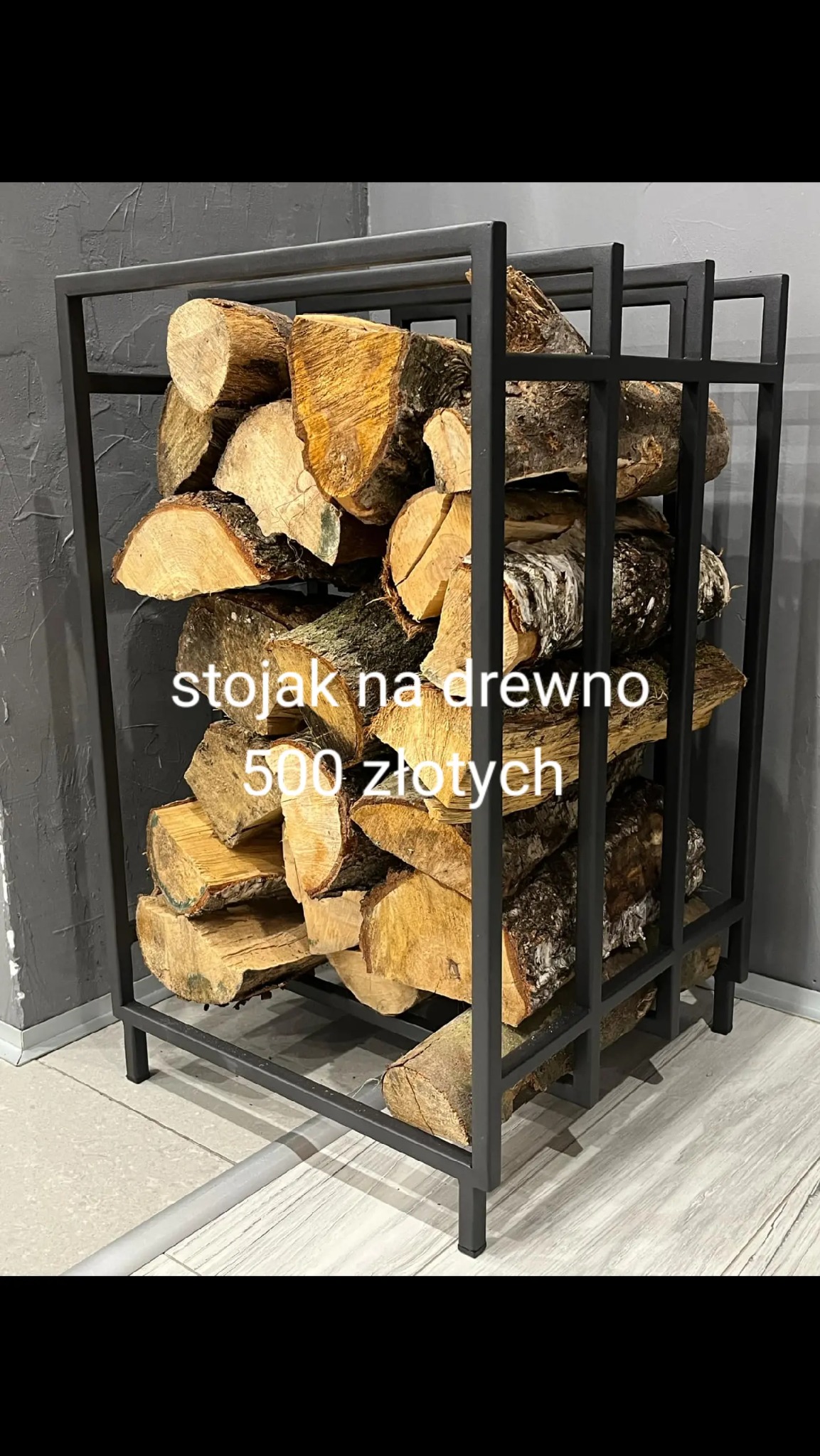 Metalowy stojak na drewno kominkowe, wypełniony pociętymi kawałkami drewna, ustawiony na jasnej podłodze w pomieszczeniu z szarą ścianą.