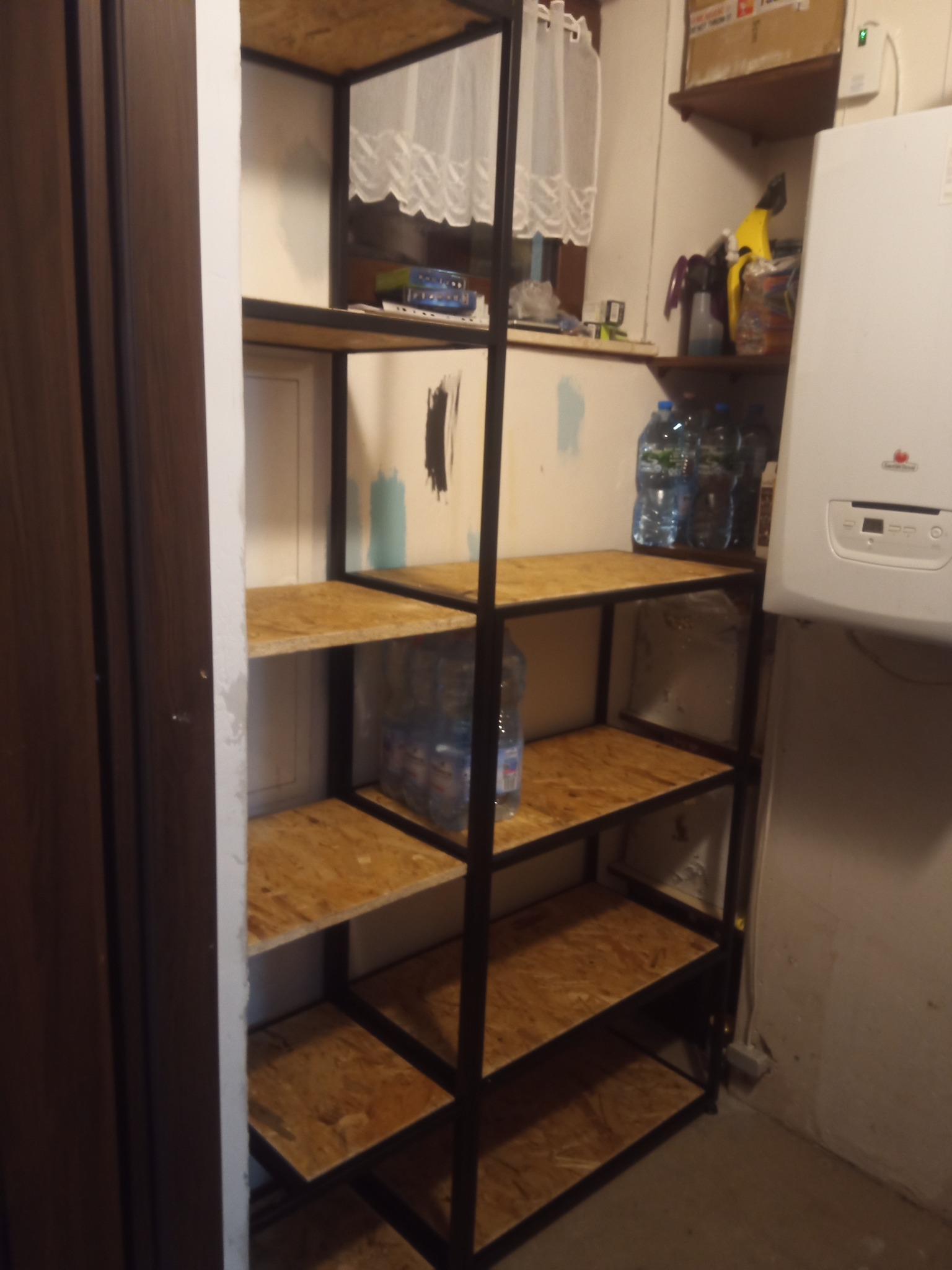 Metalowa półka z drewnianymi blatami wypełniona butelkami wody, umieszczona w pomieszczeniu gospodarczym z bojlerem i zasłoniętym oknem.