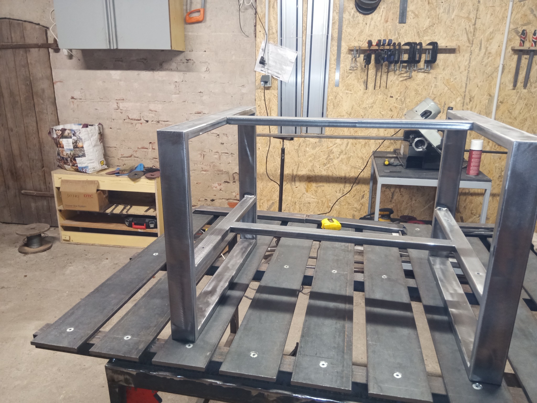 Metalowy szkielet stolika kawowego spawany na metalowym stole warsztatowym, widoczne narzędzia i akcesoria w tle.