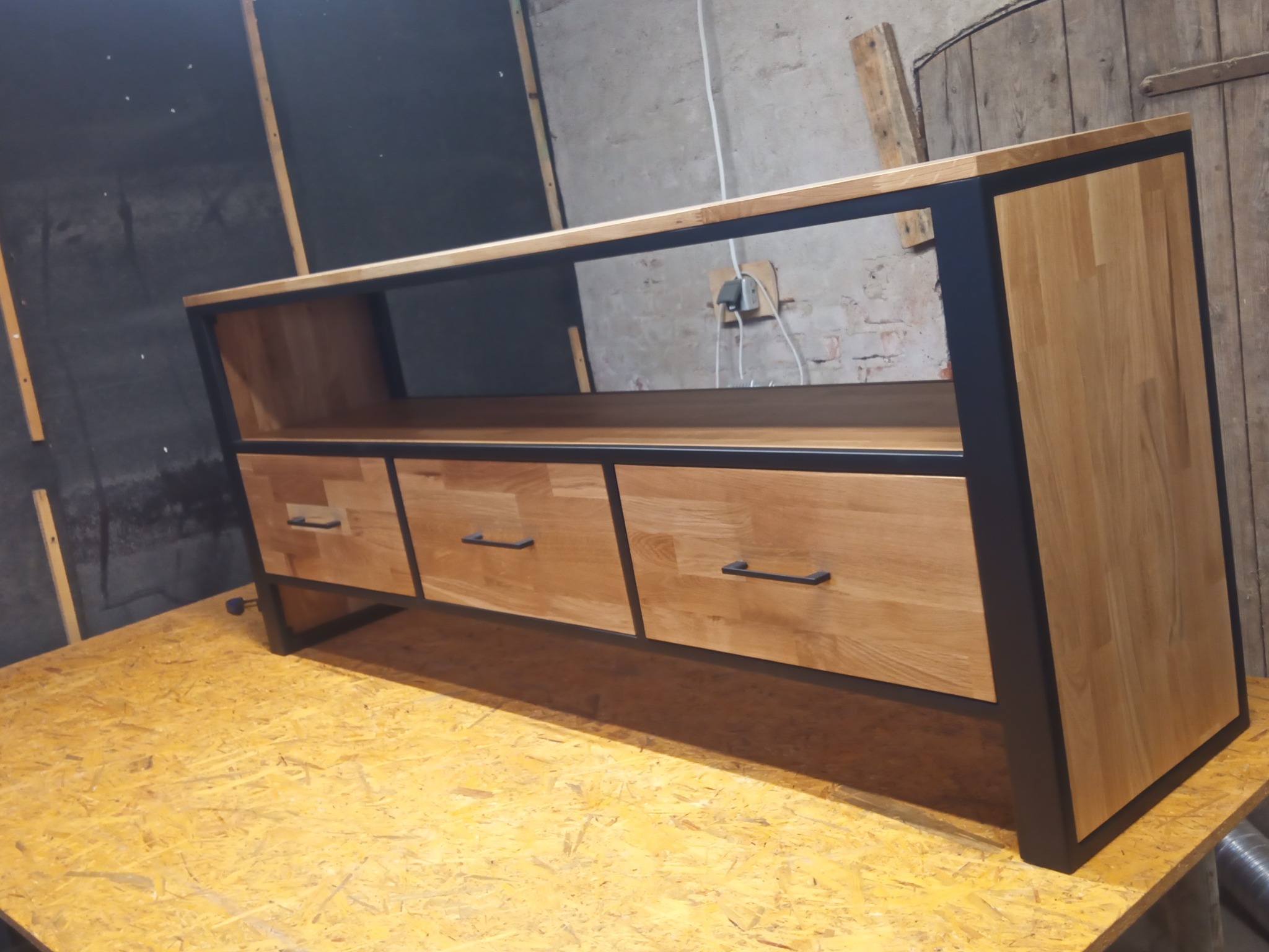 Drewniana szafka RTV z trzema szufladami i czarną ramą, wykonana na wymiar w Świerznie, stoi na płycie OSB w warsztacie.