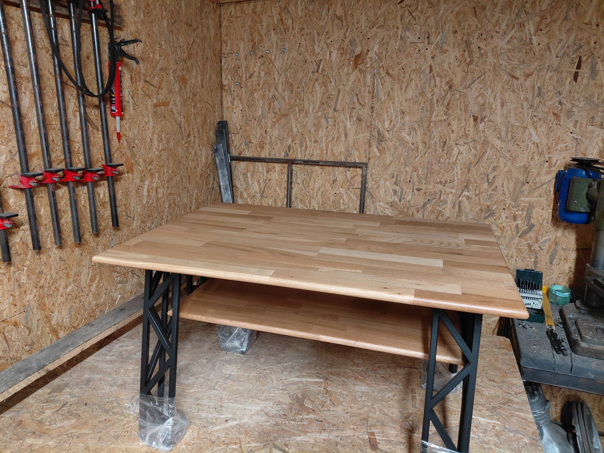 Drewniany stół z metalowymi nogami w warsztacie, nogi zabezpieczone folią. Na ścianie narzędzia i płyta OSB. Blat z jasnego drewna, widoczna półka pod blatem.