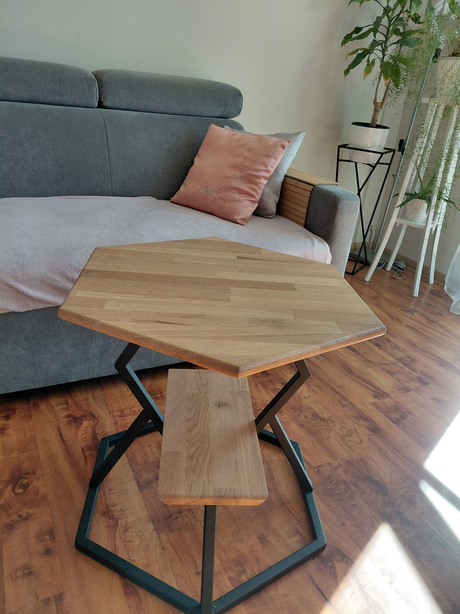 Stolik kawowy o heksagonalnym blacie z jasnego drewna i czarną, geometryczną podstawą, ustawiony w salonie obok szarej sofy z poduszką.
