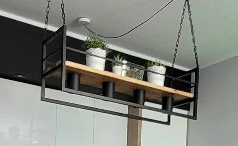 Metalowa, wisząca półka z drewnianym blatem, na której stoją doniczki z roślinami. Półka zawieszona na łańcuchach, widoczna na tle białych szafek i szarej ściany.