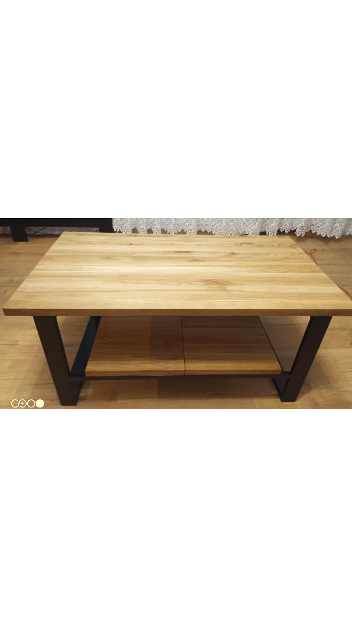 Stolik kawowy z jasnego drewna z półką, na czarnych, metalowych nogach w kształcie litery V, na drewnianej podłodze. W tle firanka.