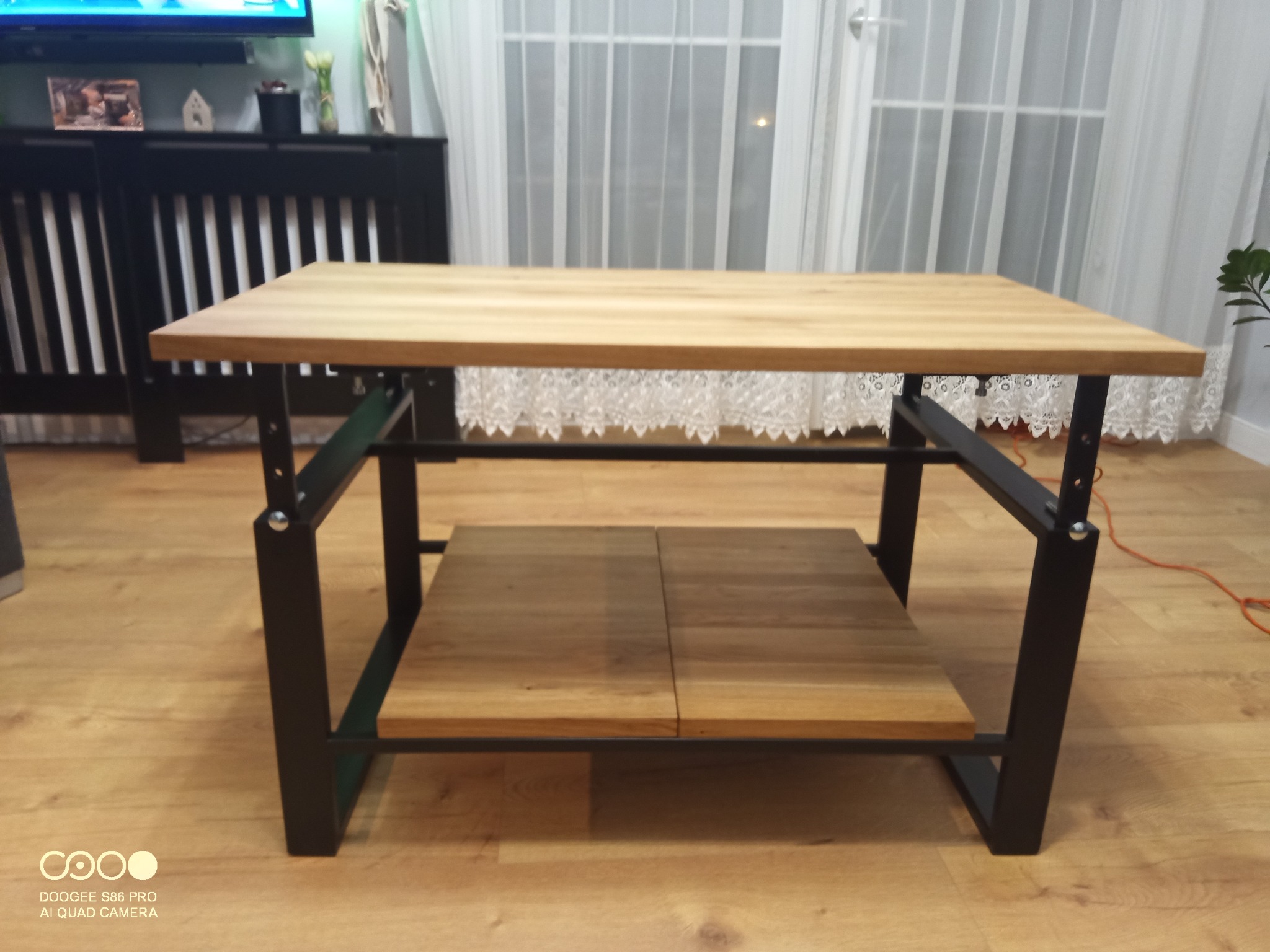 Stolik kawowy z jasnego drewna na czarnych, metalowych nogach, z dodatkową półką. W tle zasłony i komoda. Minimalistyczny design.