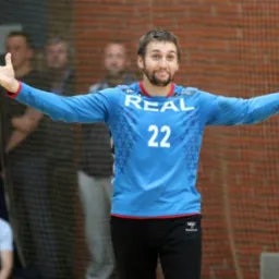 Uśmiechnięty mężczyzna w błękitnej koszulce z numerem 22, rozkłada ręce na tle siatki i obserwujących go osób. Ujęcie z dołu, oświetlenie naturalne.