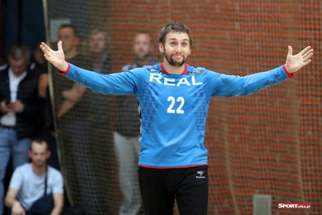 Uśmiechnięty mężczyzna w błękitnej koszulce z numerem 22, rozkłada ręce na tle siatki i obserwujących go osób. Ujęcie z dołu, oświetlenie naturalne.