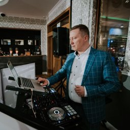 Usługi Muzyczne Michał Kluczyński - Mężczyzna w niebieskiej marynarce w kratę obsługuje sprzęt DJ-ski z laptopem na tle wzorzystej tapety w sali bankietowej.