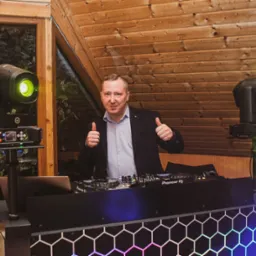 Uśmiechnięty DJ w marynarce, z podniesionymi kciukami, za konsoletą Pioneer DJ, z oświetleniem scenicznym w tle.