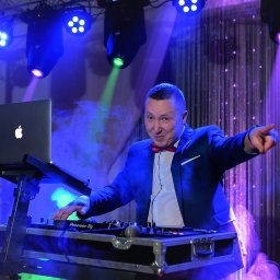 Usługi Muzyczne Michał Kluczyński - Uśmiechnięty DJ w niebieskiej marynarce i czerwonej muszce, wskazujący palcem, grający na sprzęcie Pioneer DJ pod wielobarwnym oświetleniem scenicznym i z laptopem Apple.