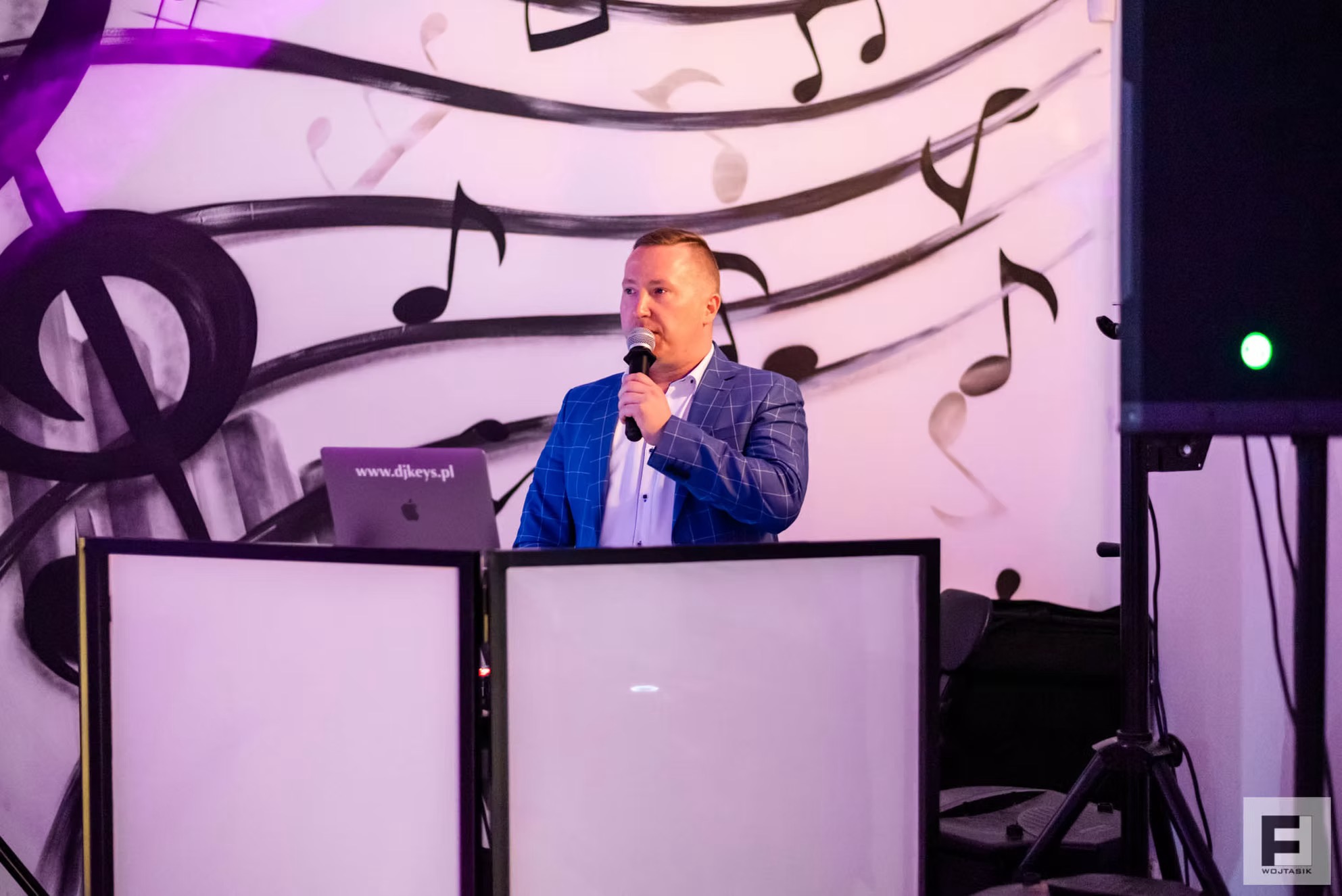 Prezenter w niebieskiej marynarce, trzymający mikrofon, za konsoletą DJ-ską z laptopem i tłem z motywem nut.