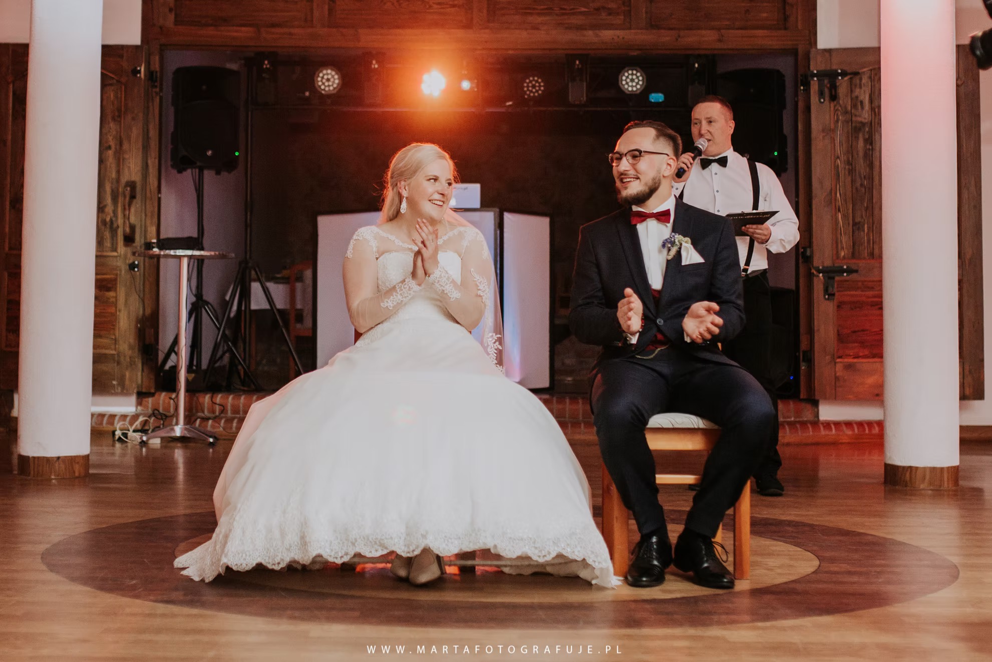 Para młoda siedzi na krzesłach i klaszcze, za nimi konferansjer z mikrofonem i oświetlenie sceniczne, drewniane drzwi w tle.