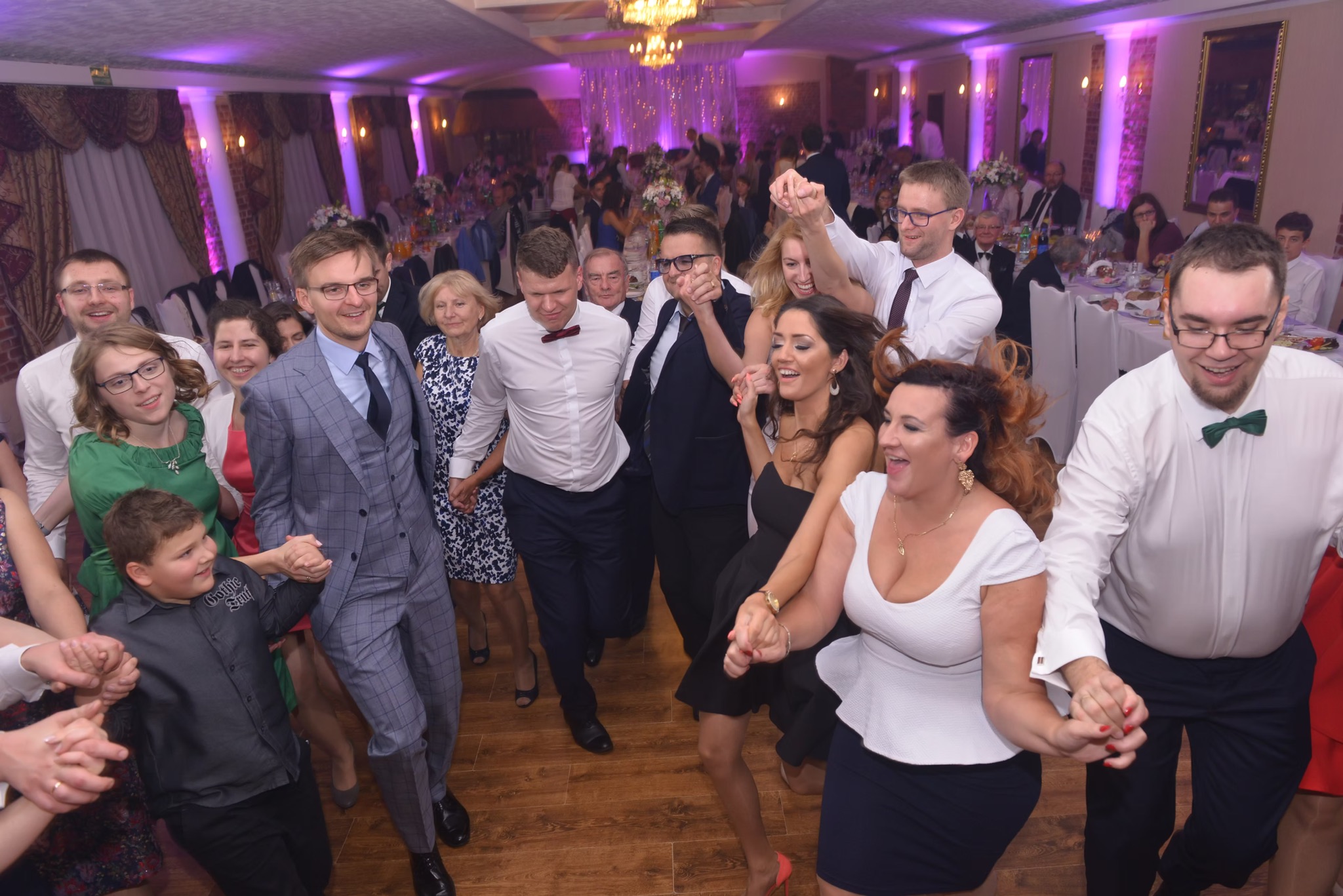 Roztańczona grupa gości weselnych w sali bankietowej z parkietem, oświetlona fioletowymi światłami, podczas tradycyjnego korowodu.