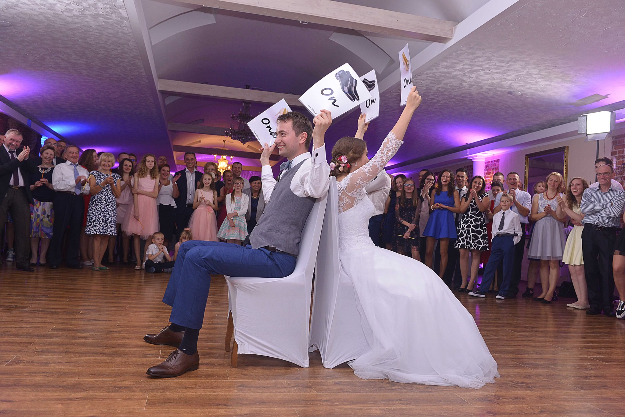 Para młoda siedzi na krzesłach tyłem do siebie, trzymając w górze kartki z napisem 'On' i 'Ona' podczas zabawy weselnej, w tle zgromadzeni goście klaszczą.
