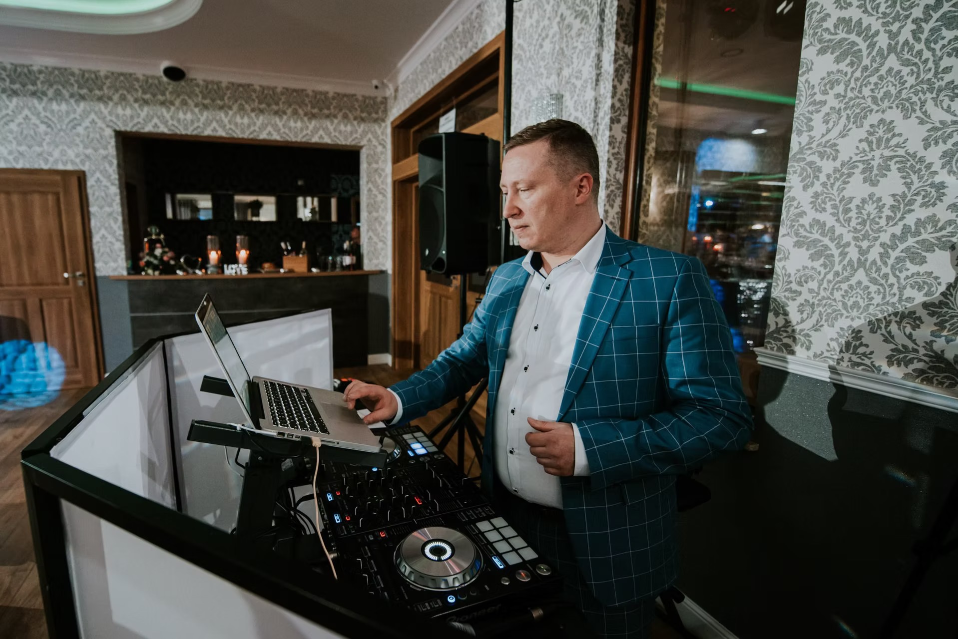 Mężczyzna w niebieskiej marynarce w kratę obsługuje sprzęt DJ-ski z laptopem na tle wzorzystej tapety w sali bankietowej.