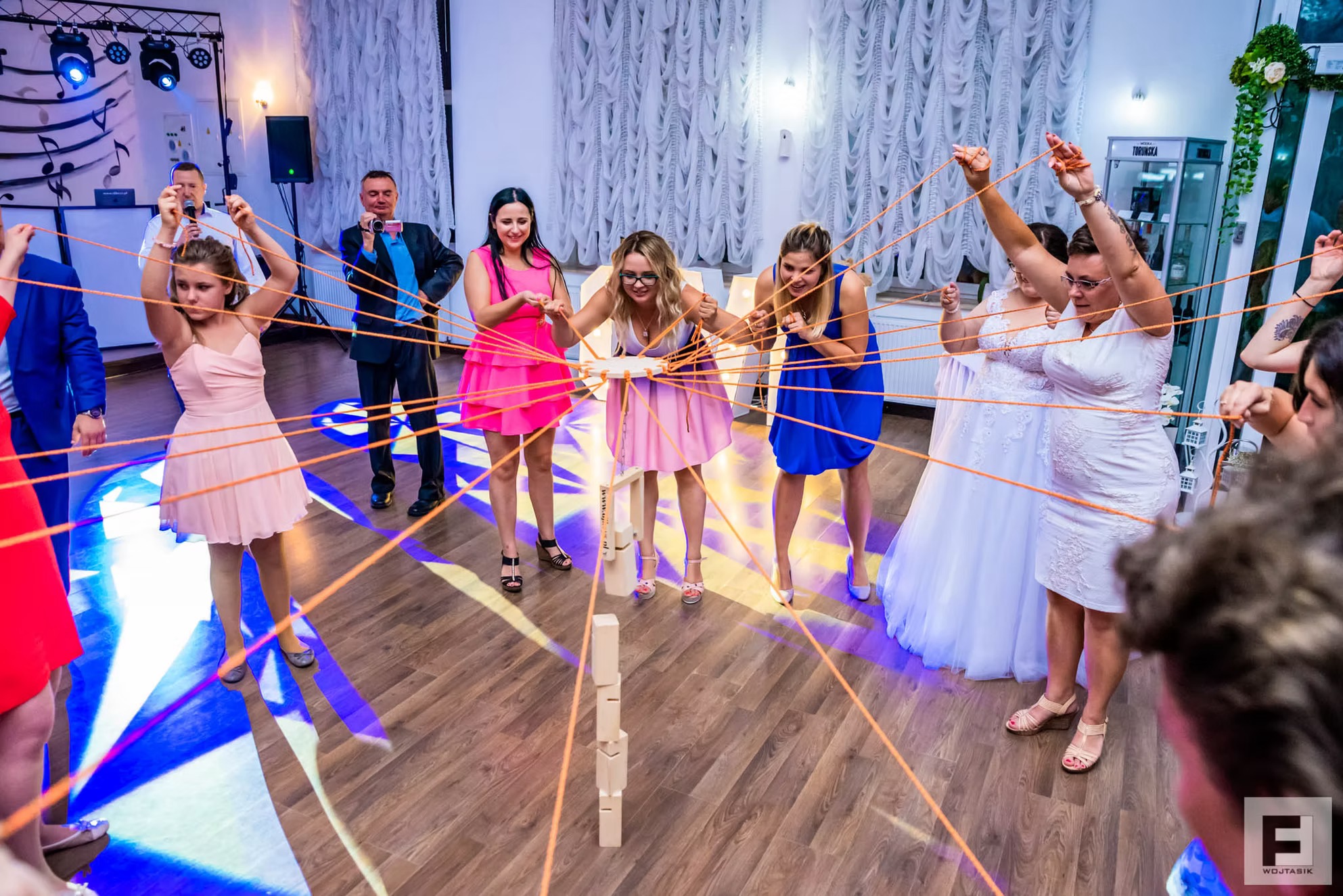 Goście weselni w kolorowych sukienkach grają w grę z drewnianymi klockami i sznurkami podczas przyjęcia, oświetlenie sceniczne w tle, na podłodze wzory świetlne.