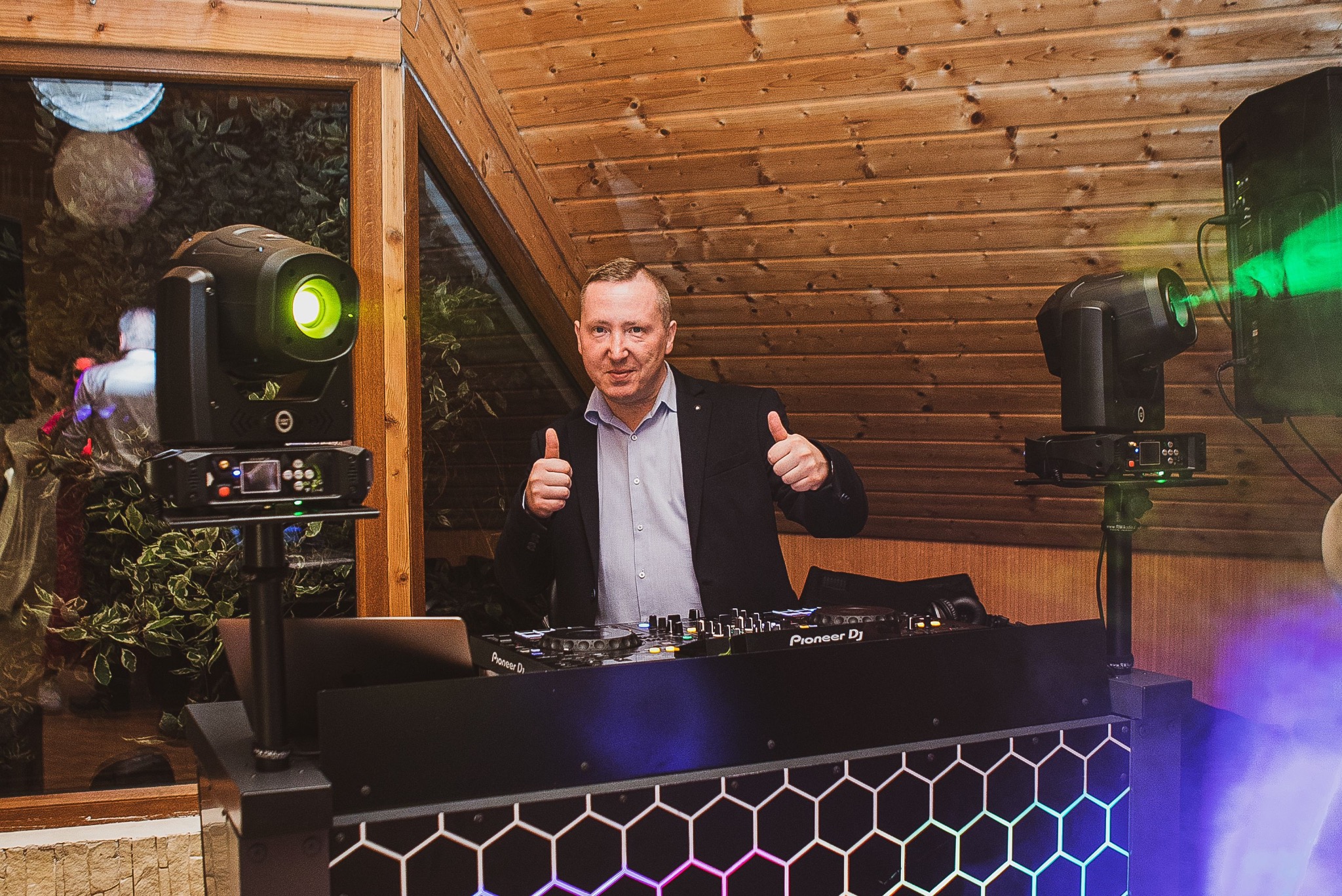 Uśmiechnięty DJ w marynarce, z podniesionymi kciukami, za konsoletą Pioneer DJ, z oświetleniem scenicznym w tle.